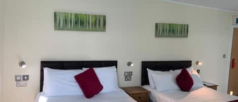 Habitación de hotel con dos camas, cada una con ropa blanca y almohadas rojas, y un cabecero oscuro. Hay dos lámparas de pared a cada lado de las camas.