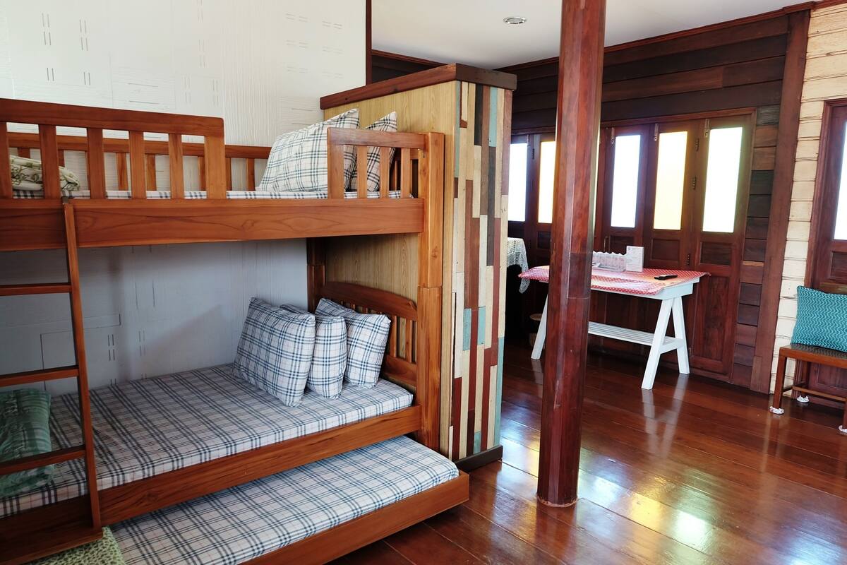 A bunk bed with plaid bedding, a wooden desk, and a chair in a room with wooden floors and walls.