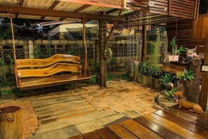 Een houten bankje onder een houten pergola, omringd door potplanten en een kleine hondbeeld.