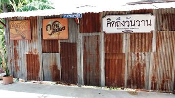 A corrugated metal wall with signs for Coca-Cola, a local brand, and a homestay named Baanchompoon Homestay.