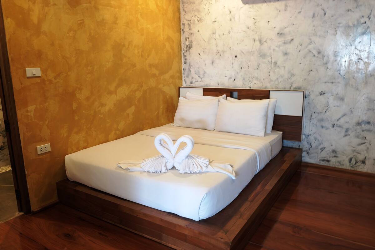 A neatly made bed with white linens and a heart-shaped towel arrangement, a wooden headboard, and a marble-patterned wall.