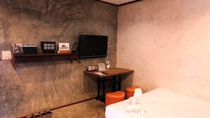 A hotel room with a bed, a desk with a flat-screen TV, a shelf with branded items, and a pair of slippers. - BaanChompooAmphawa (Amphawa)