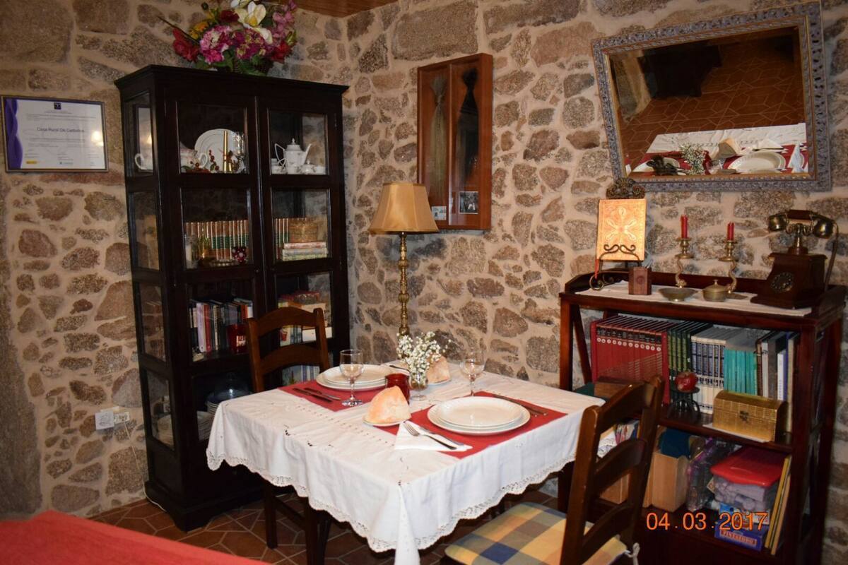 Un coin repas avec un mur en pierre, un buffet en bois doté de portes en verre, une table dressée pour deux personnes et un miroir placé au-dessus de la table.