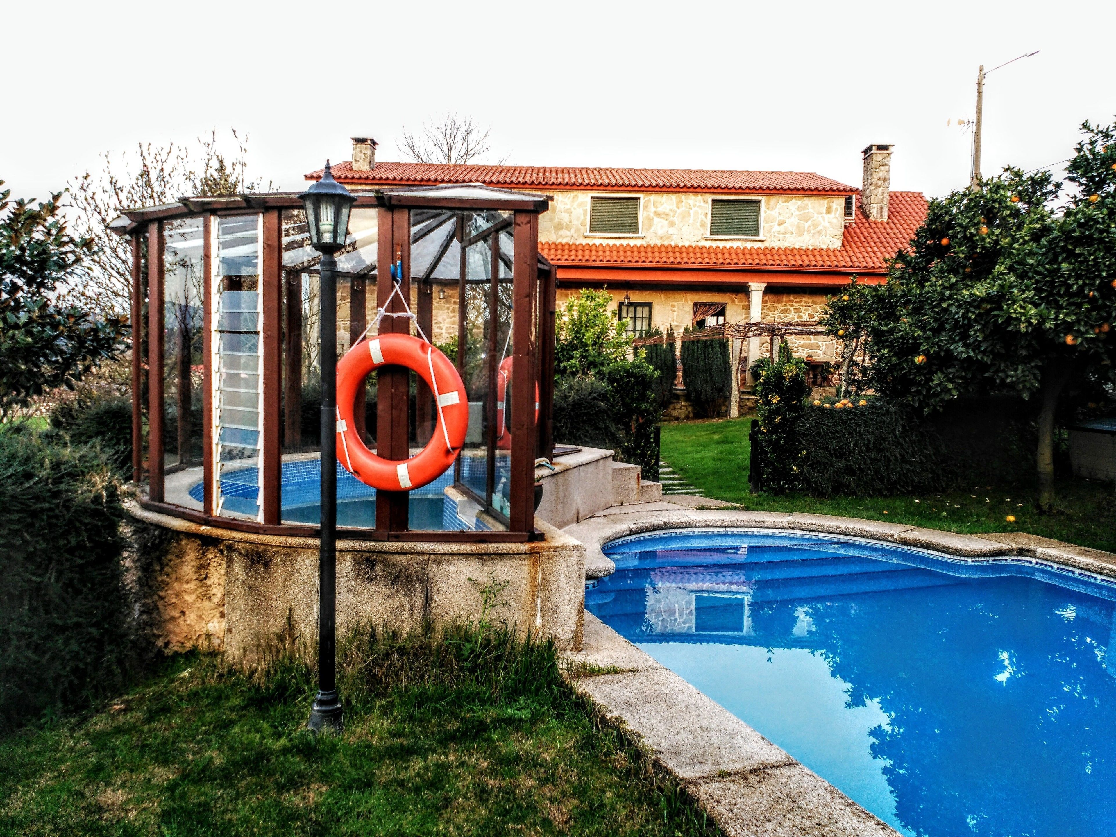 Ein Schwimmbecken mit Glasüberdachung und Rettungsboje, umgeben von Grünpflanzen und einem dahinterliegenden Steinhaus.