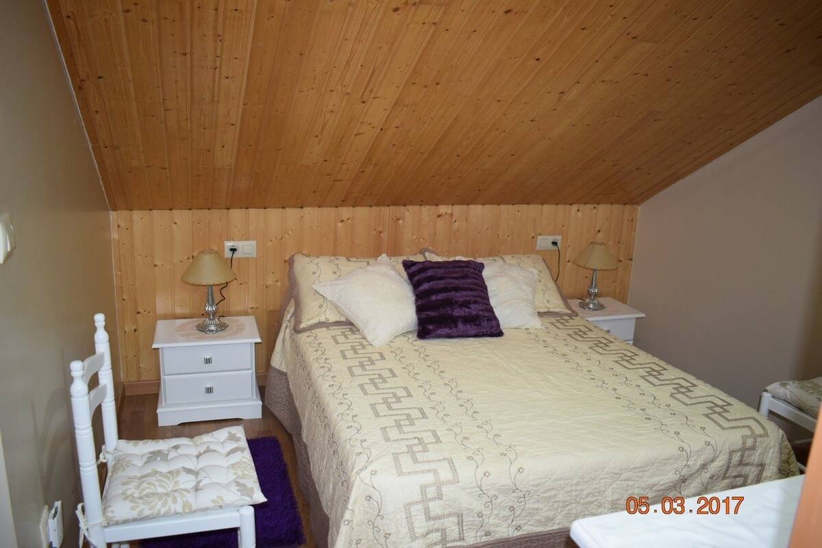 Une chambre avec un plafond en bois, un lit avec une literie à motifs, deux tables de chevet avec des lampes et une chaise avec un coussin.