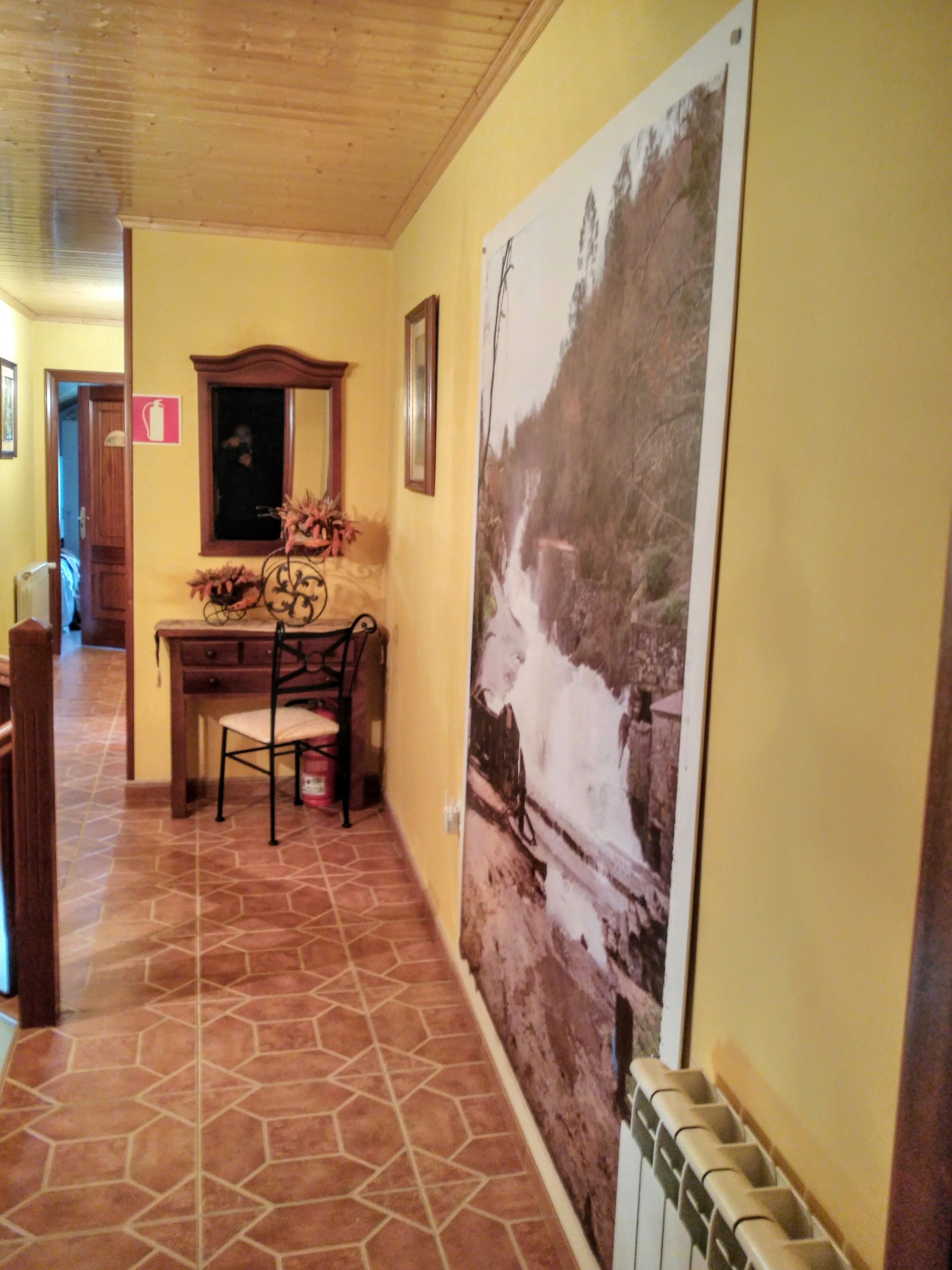A hallway with a tiled floor, a wooden desk with a chair, a large framed picture on the wall, and a radiator.