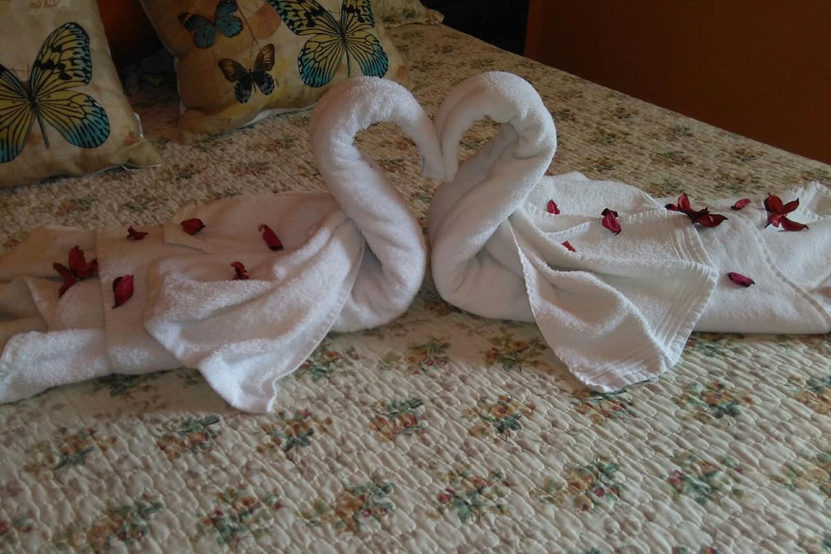 Un lit recouvert d’une courtepointe à motifs floraux et agrémenté de deux serviettes blanches disposées en forme de cœur, avec des pétales de rose.