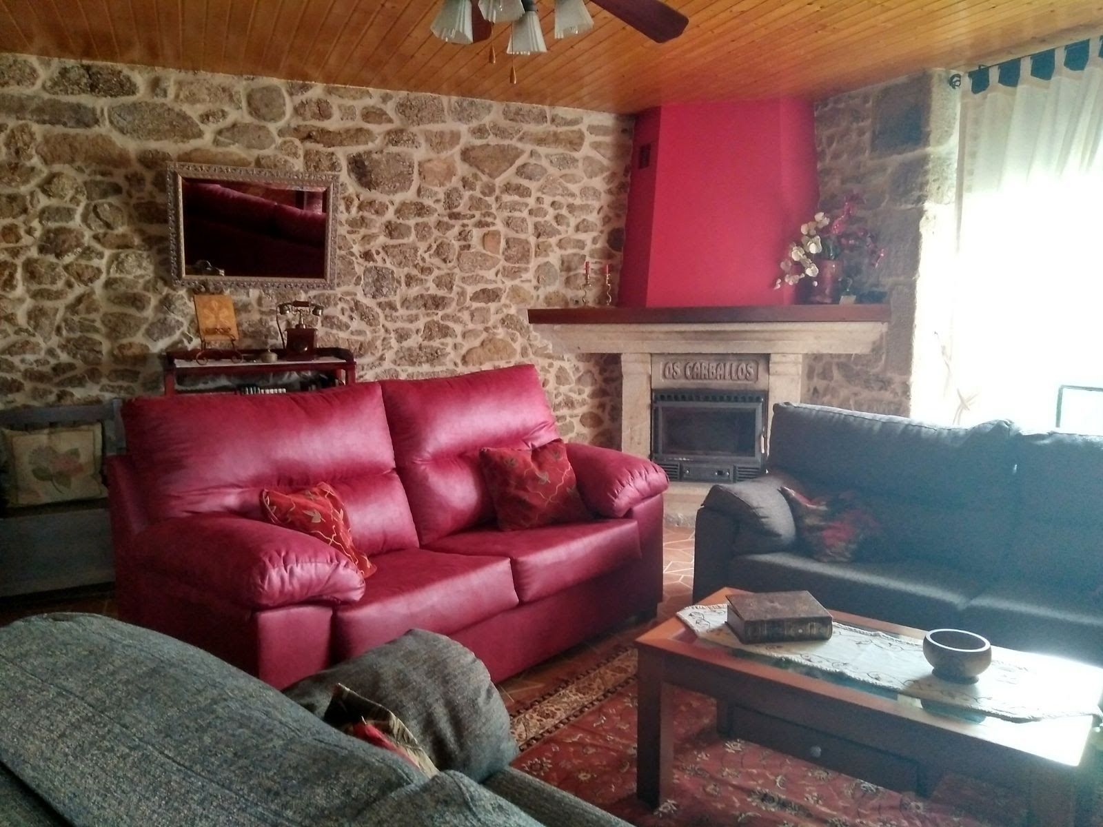 A cozy living room with a stone wall, a fireplace labeled "OS CARBALOS," a red leather sofa, and a wooden coffee table.
