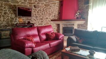 A cozy living room with a stone wall, a fireplace labeled "OS CARBALOS," a red leather sofa, and a wooden coffee table.