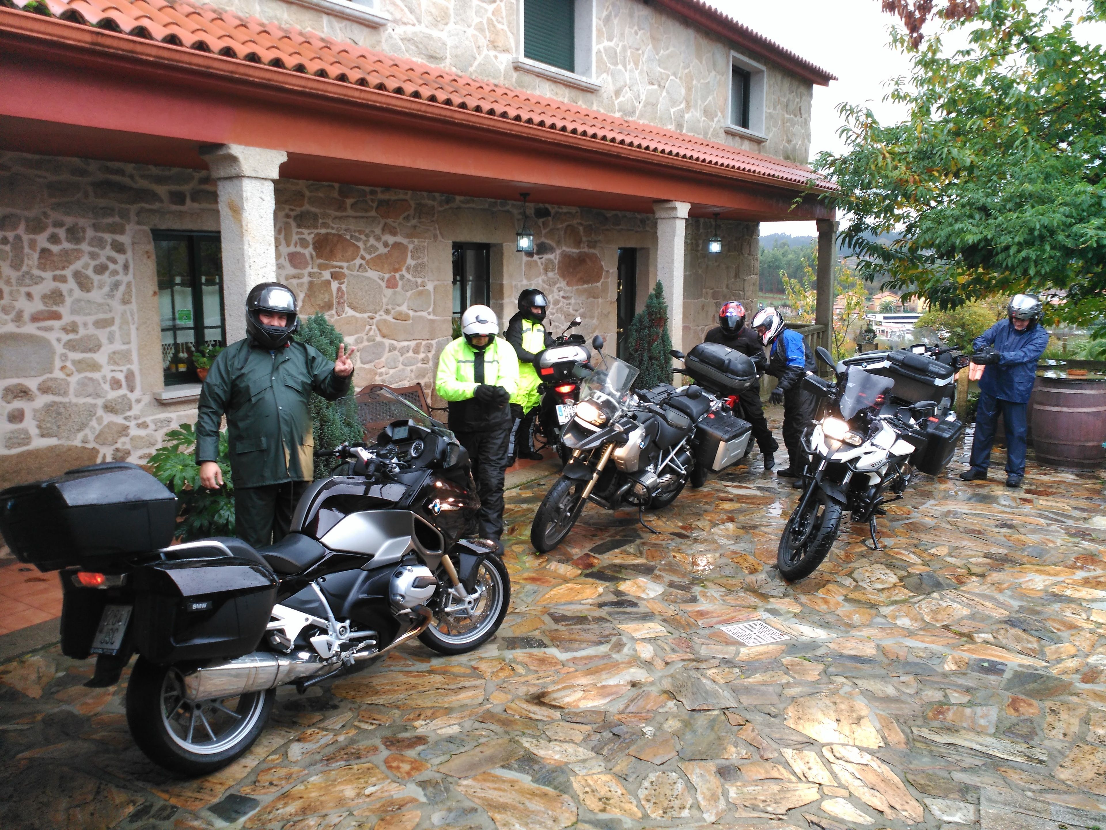 A stone building with a red-tiled roof, parked motorcycles, and a person in a blue jacket standing by a barrel.