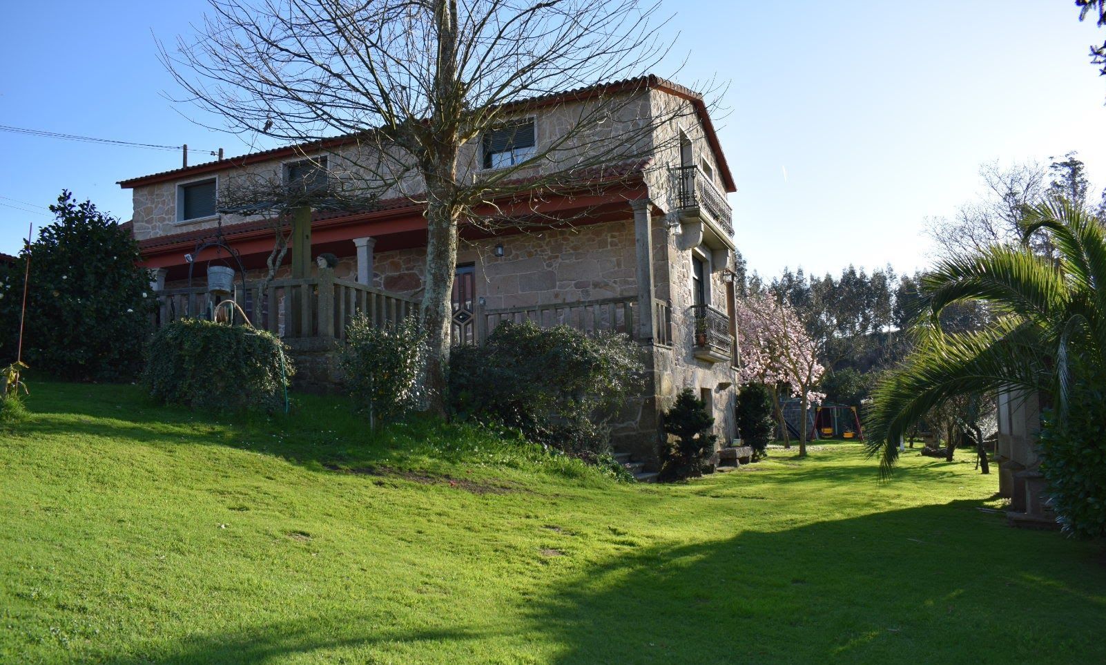 Uma casa de pedra de dois andares com telhado vermelho, cercada por um gramado bem cuidado e várias árvores.