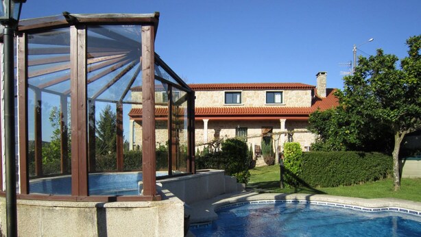 A swimming pool with a glass enclosure, a stone building with a red roof, and a well-maintained garden. - Casa Rural Os Carballos (Barro)