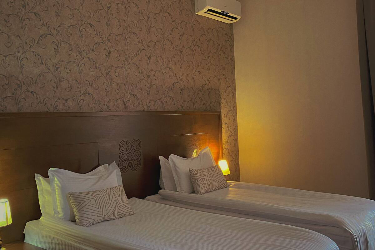 A hotel room with two beds, a headboard with a logo, an air conditioning unit, and a nightstand with a lamp.