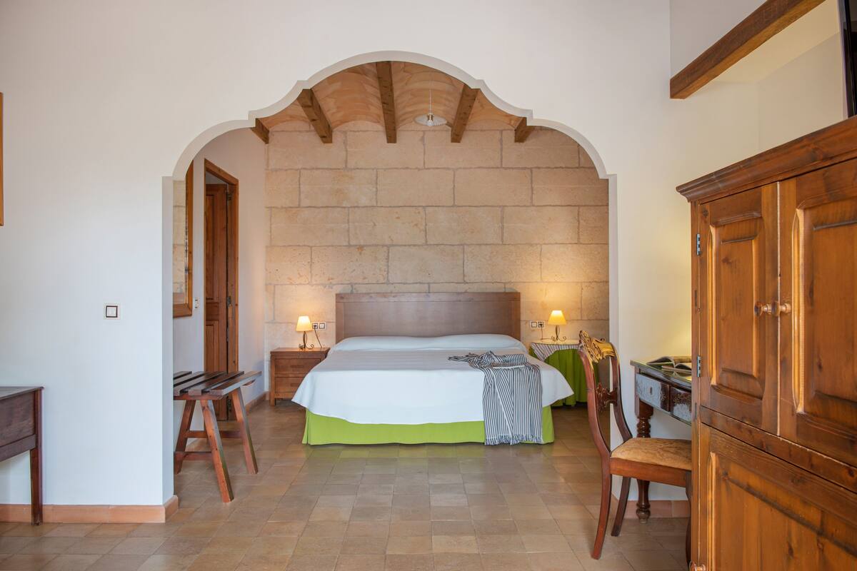 A bedroom with a large bed, wooden headboard, two bedside tables with lamps, a wooden chair, and a wooden wardrobe.
