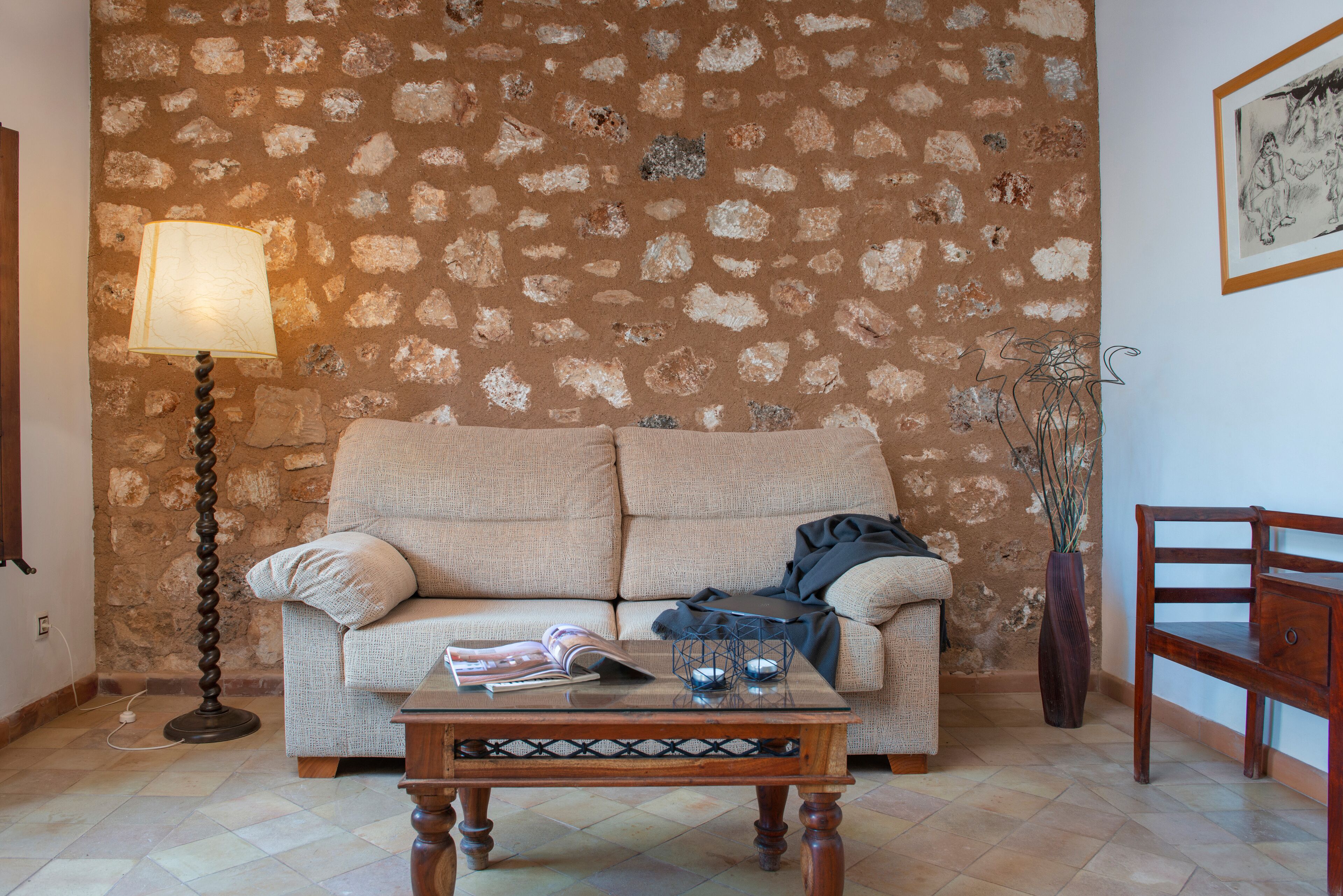 Una sala de estar con pared de piedra, un sofá, una mesa de centro con revistas, una lámpara de pie y un jarrón con plantas.