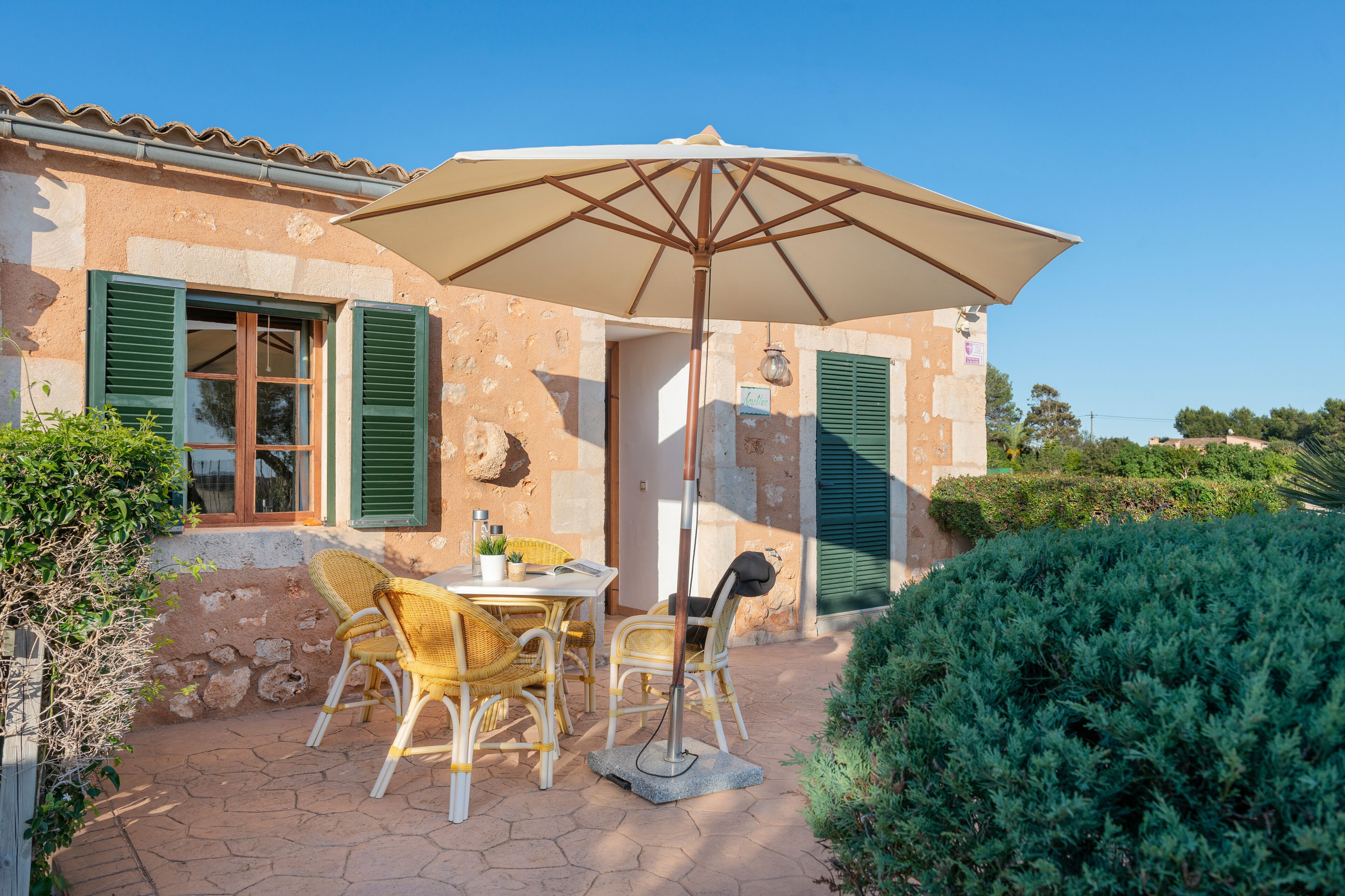 Un patio con una mesa, sillas, una sombrilla y un edificio de piedra con contraventanas verdes.