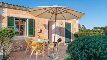 Un patio con una mesa, sillas, una sombrilla y un edificio de piedra con contraventanas verdes.