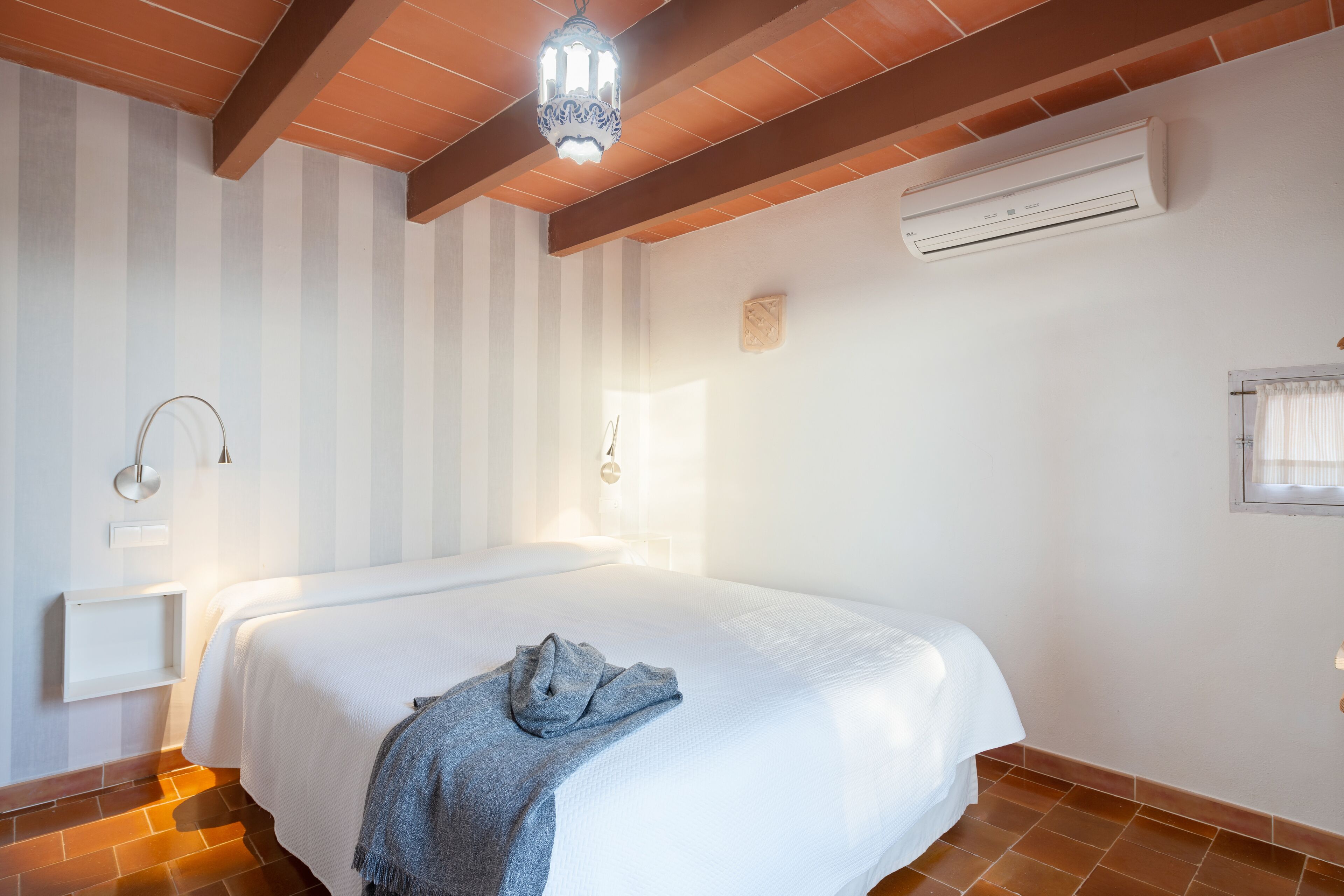A bedroom with a large bed, an air conditioning unit, a wall-mounted light fixture, and a window with curtains.
