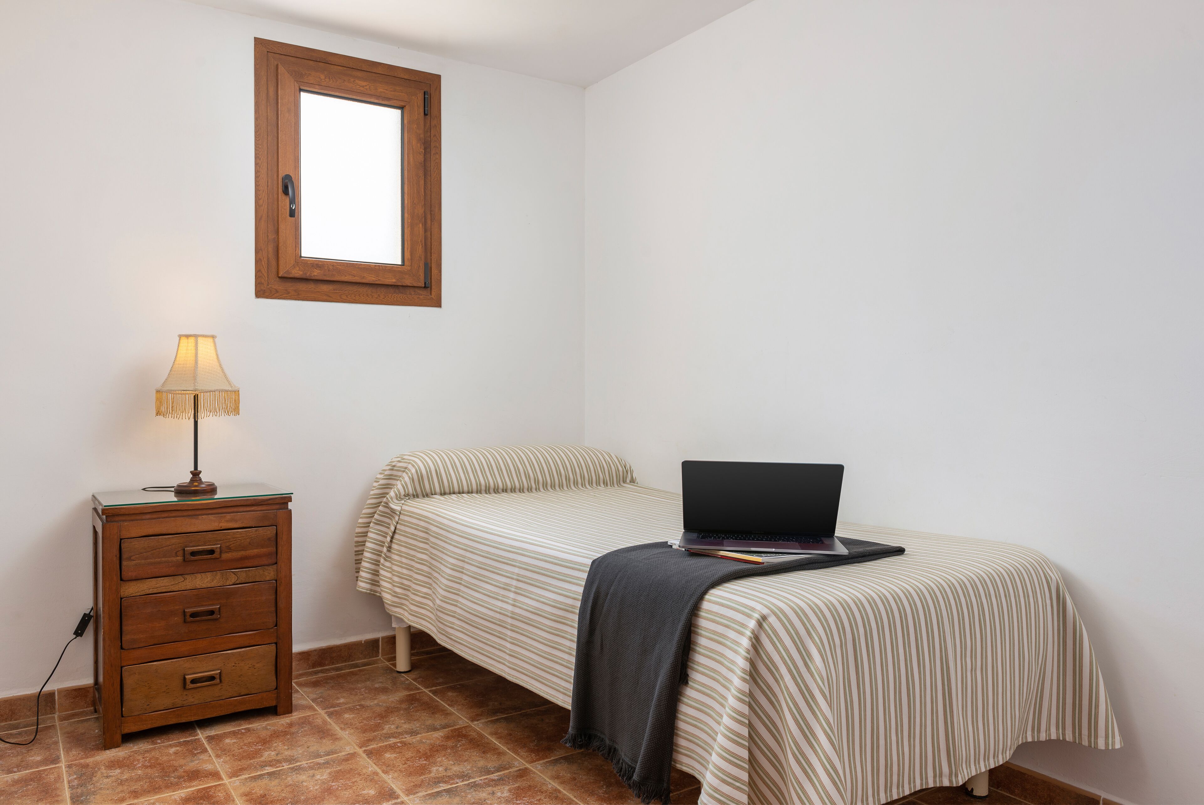 A small, neatly arranged bedroom with a single bed, a bedside table with a lamp, and a laptop on the bed.