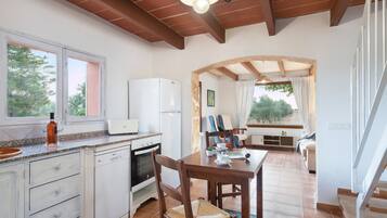 Een keuken met witte apparaten, een eethoek met een houten tafel en stoelen, en uitzicht op het landschap door grote ramen.