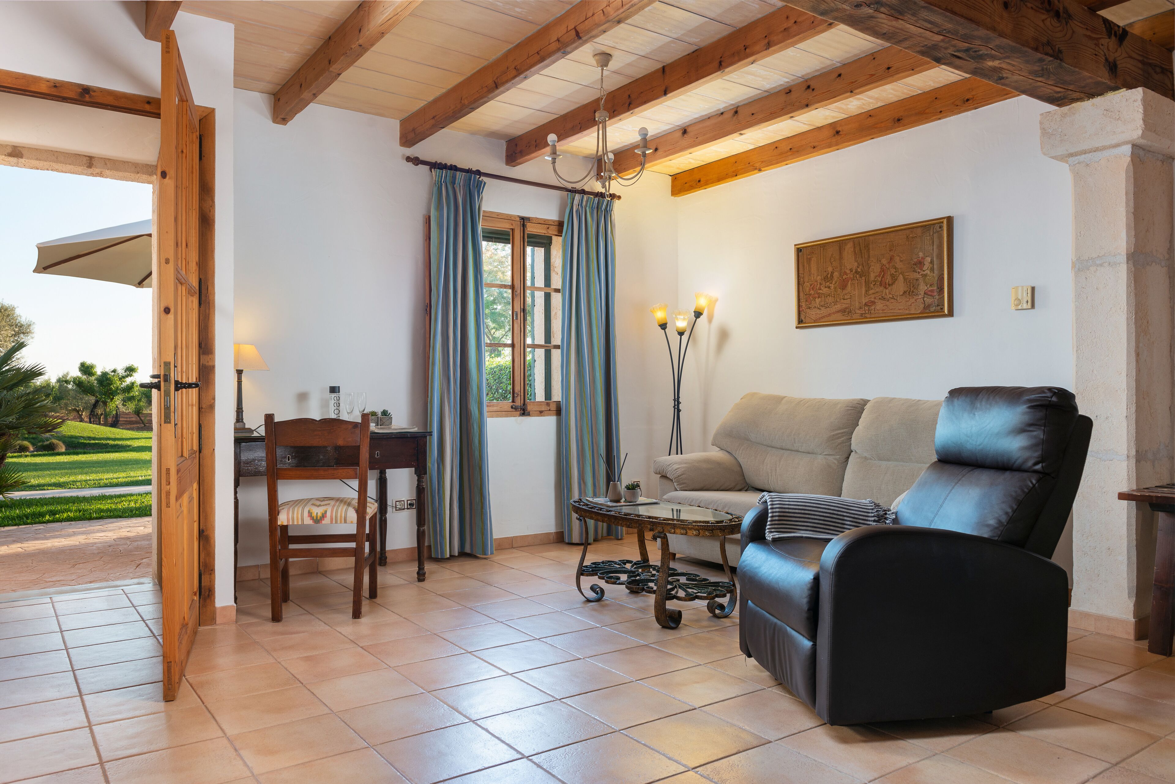 Una sala de estar con un sofá, un sillón, una mesita y una lámpara. Hay una ventana con cortinas y un cuadro en la pared.