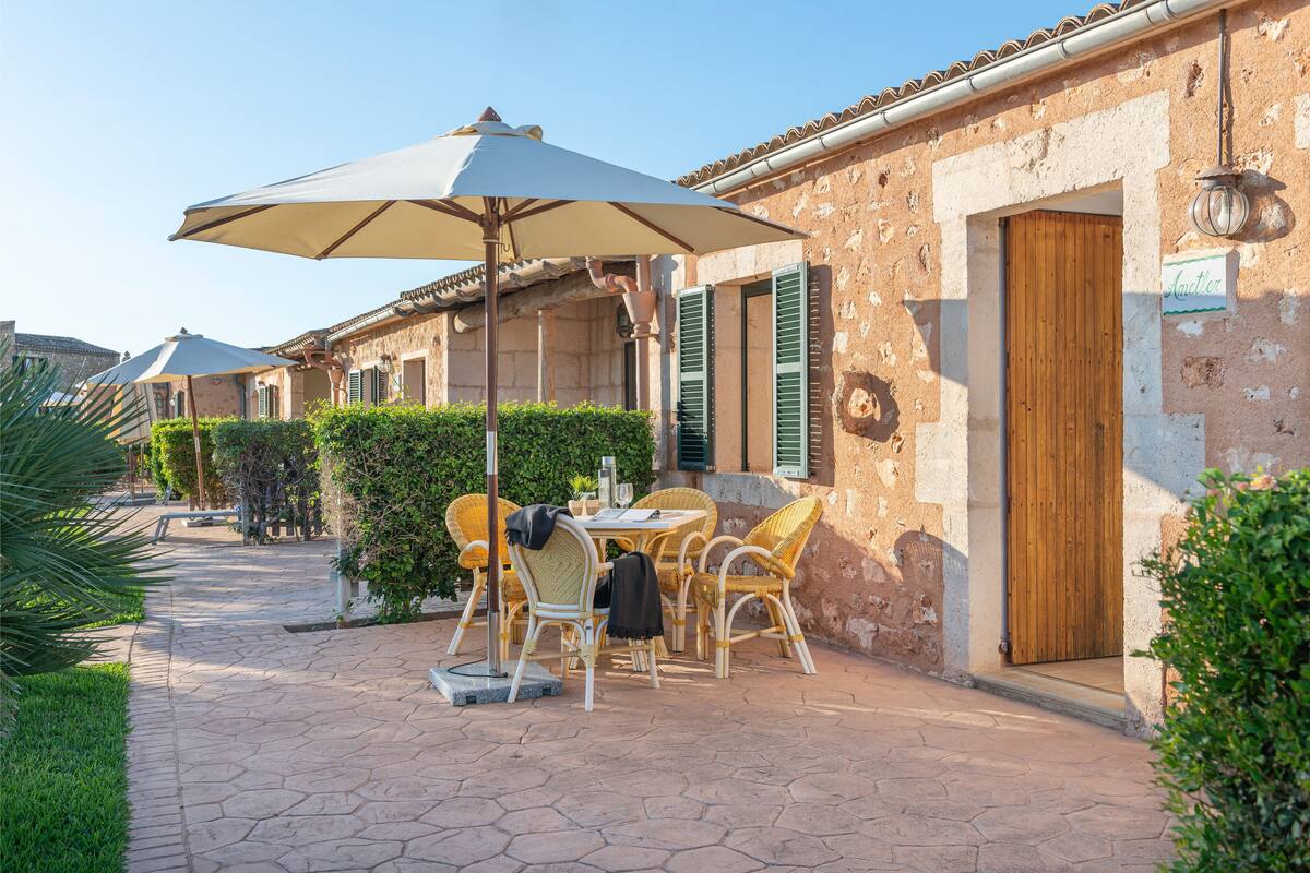 A patio with wicker furniture and a large umbrella, a stone pathway, and a building with a wooden door and green shutters.
