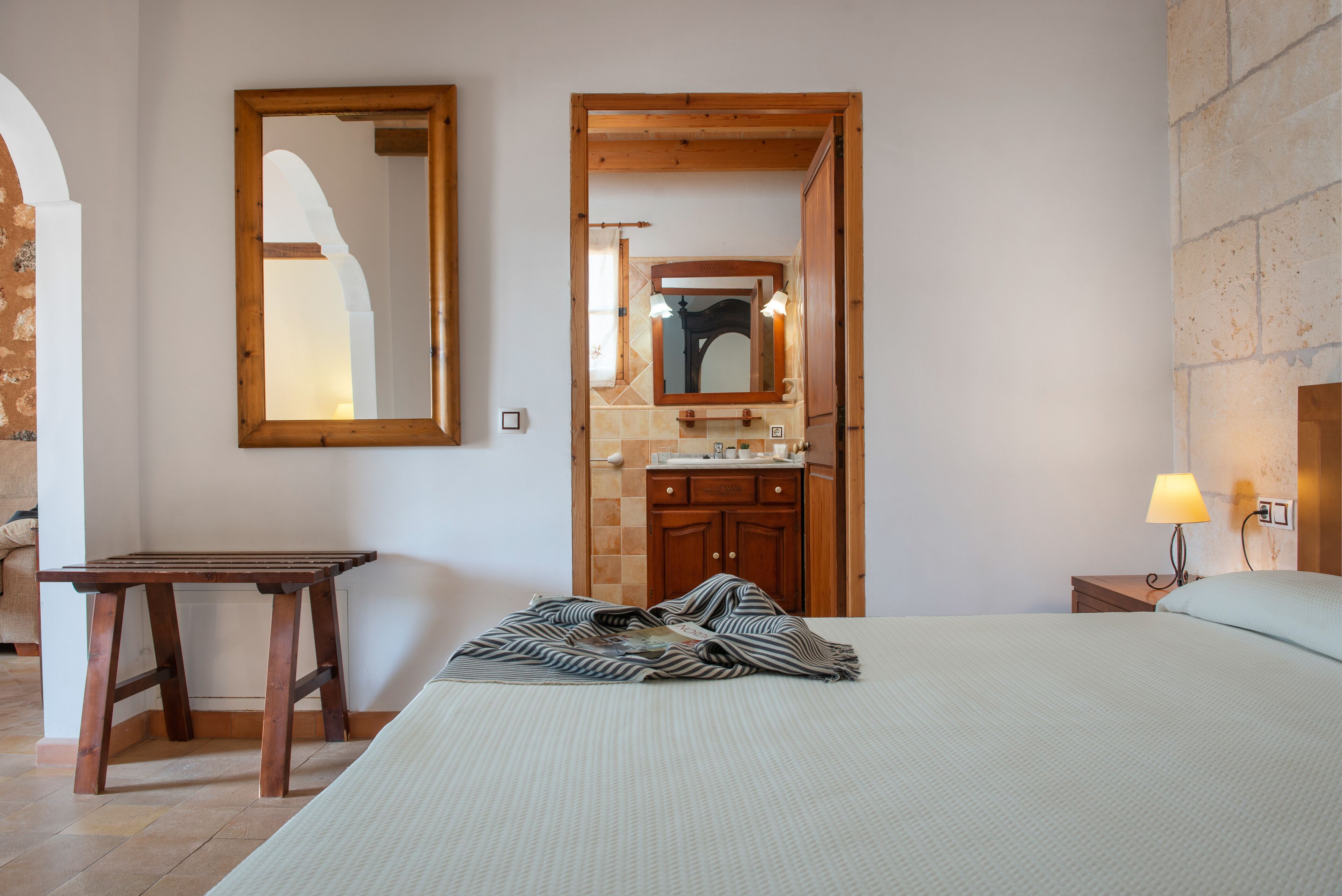 Une chambre à coucher avec un lit, une table de chevet en bois avec une lampe, une petite table et une salle de bain visible à travers une porte ouverte.