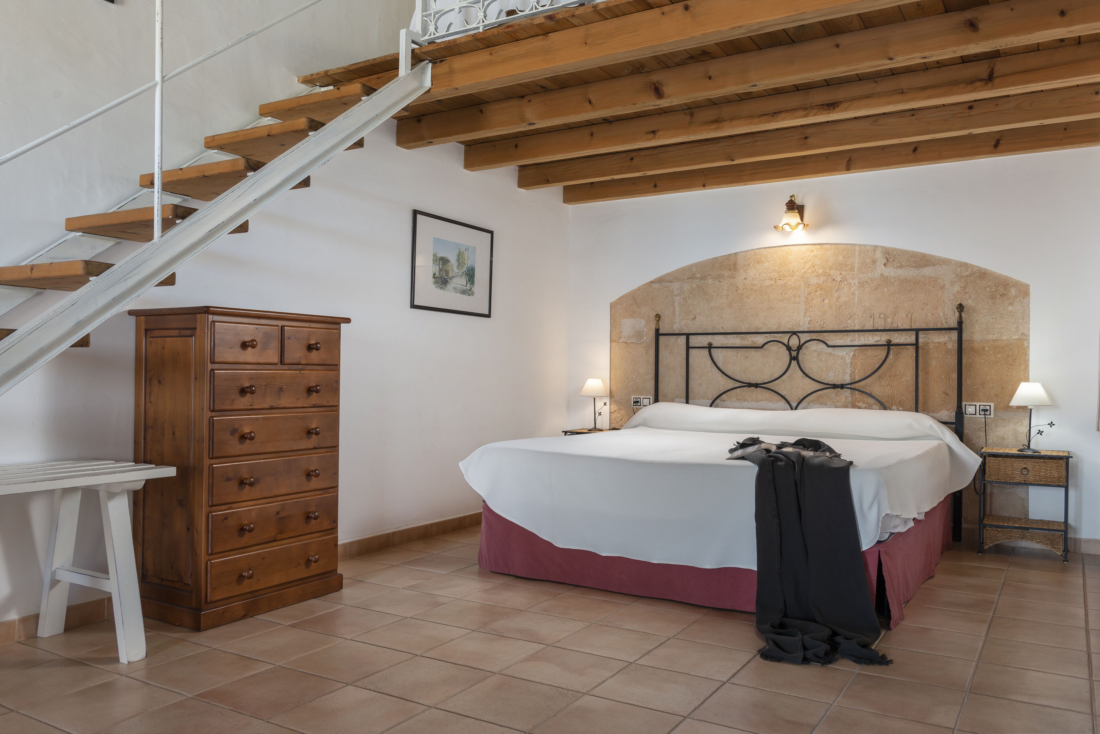 Un dormitorio con una escalera de madera, una cama con cabecera, una mesita de noche, un aparador y un cuadro en la pared.