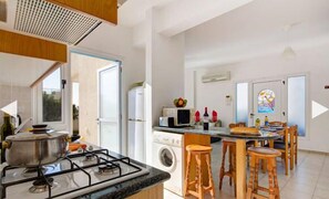 A modern kitchen with a gas stove, microwave, and washing machine. There are wooden stools and a dining table with chairs.