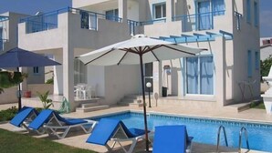 A modern building with a pool, blue lounge chairs, and umbrellas. - Philippos Villas (Chlorakas)
