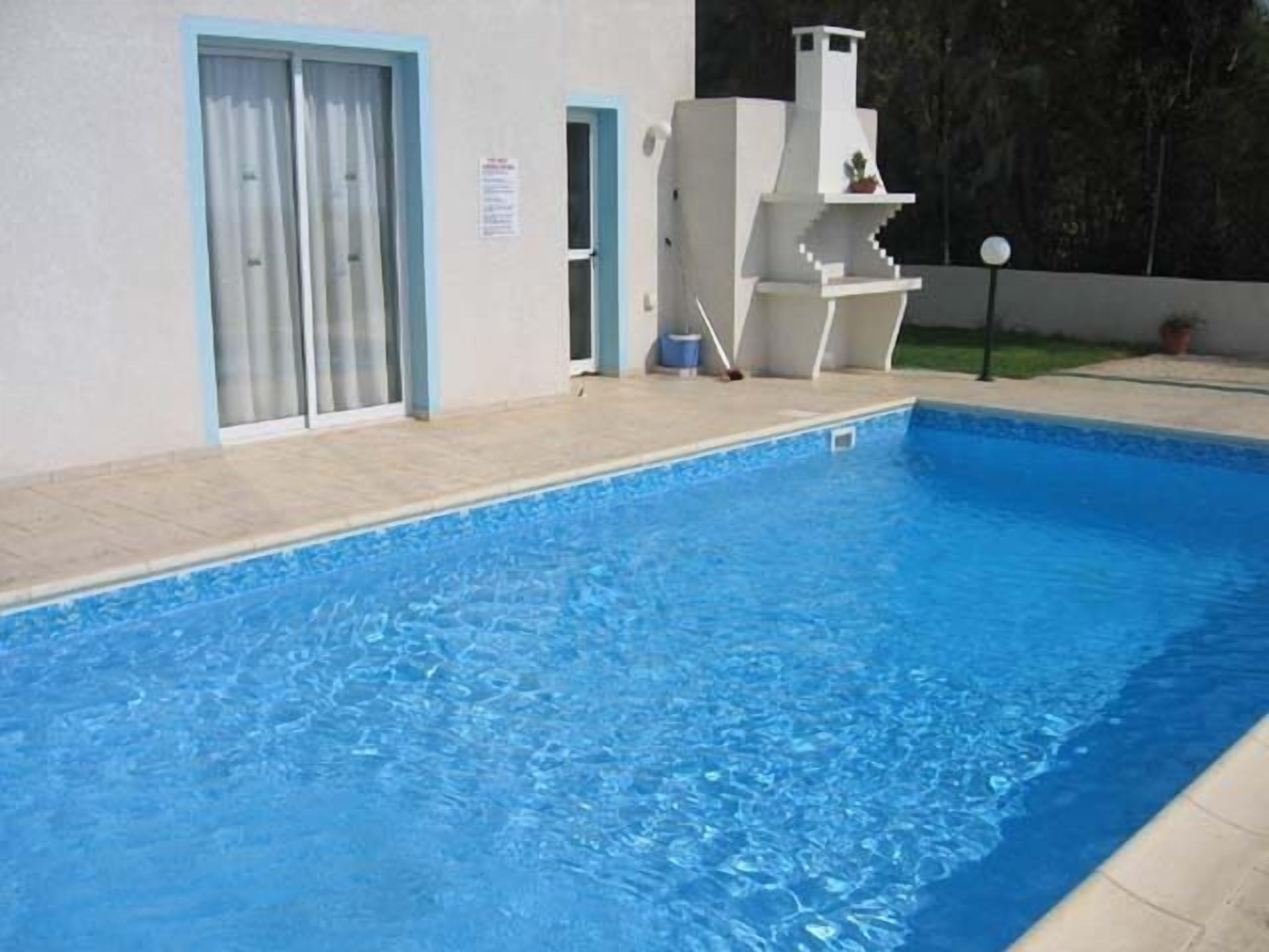 A swimming pool with a tiled deck, a white structure with steps, and a door with a blue frame.