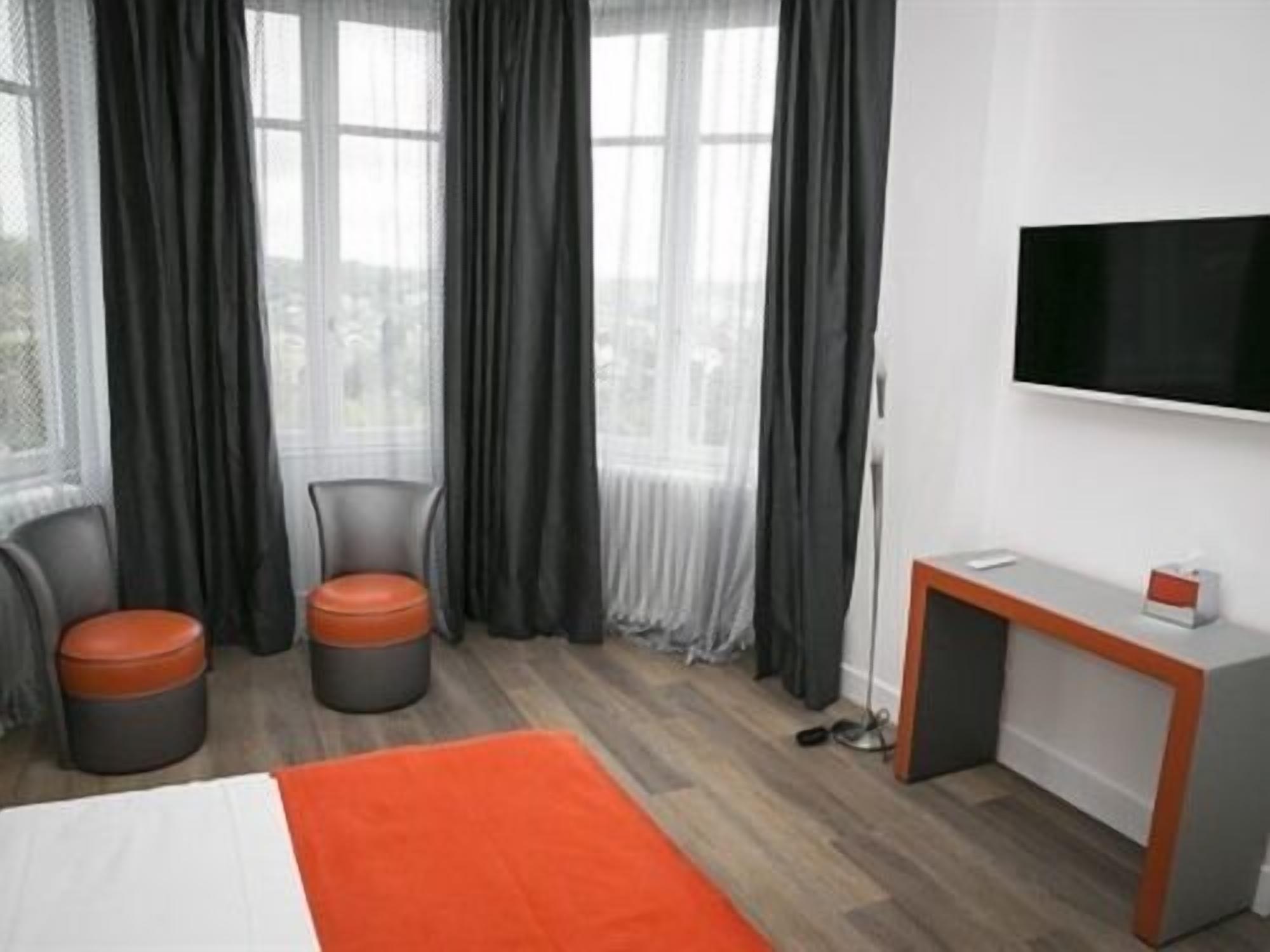 A hotel room with a bed, two chairs, a small table, a TV, and large windows with curtains.