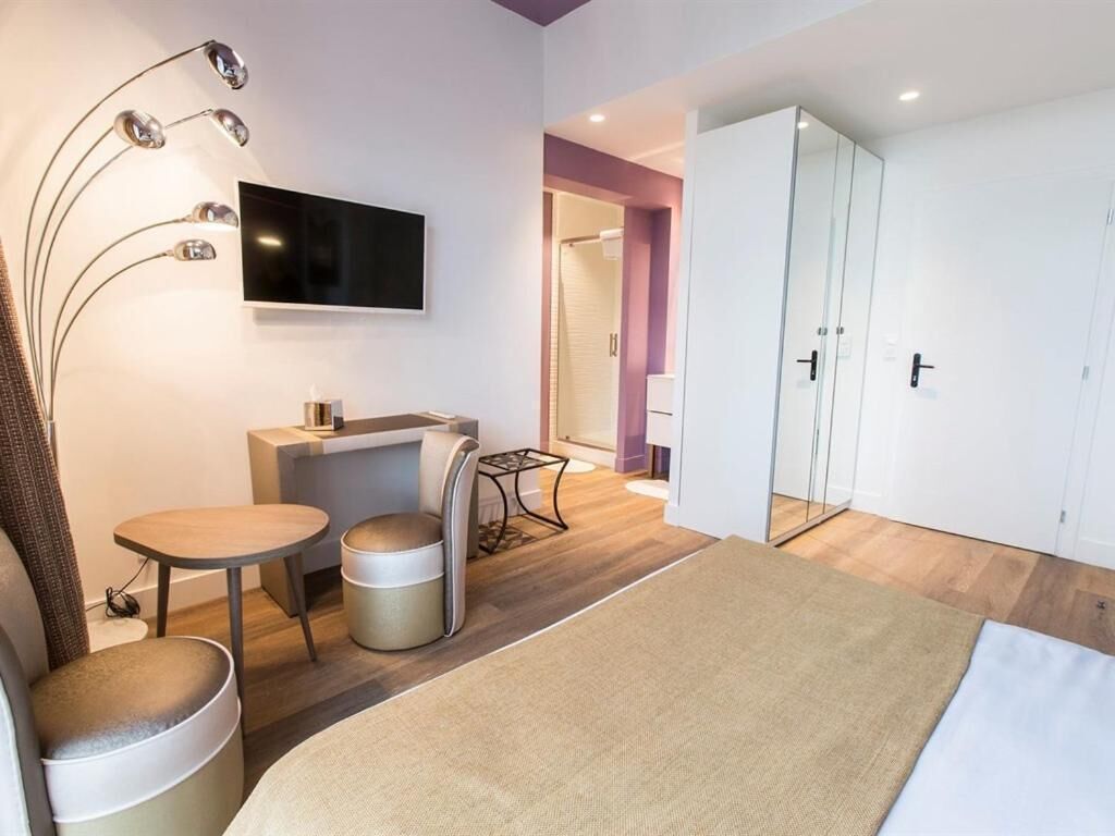 A modern hotel room with a bed, a desk, a chair, a small table, a TV, and a wardrobe.