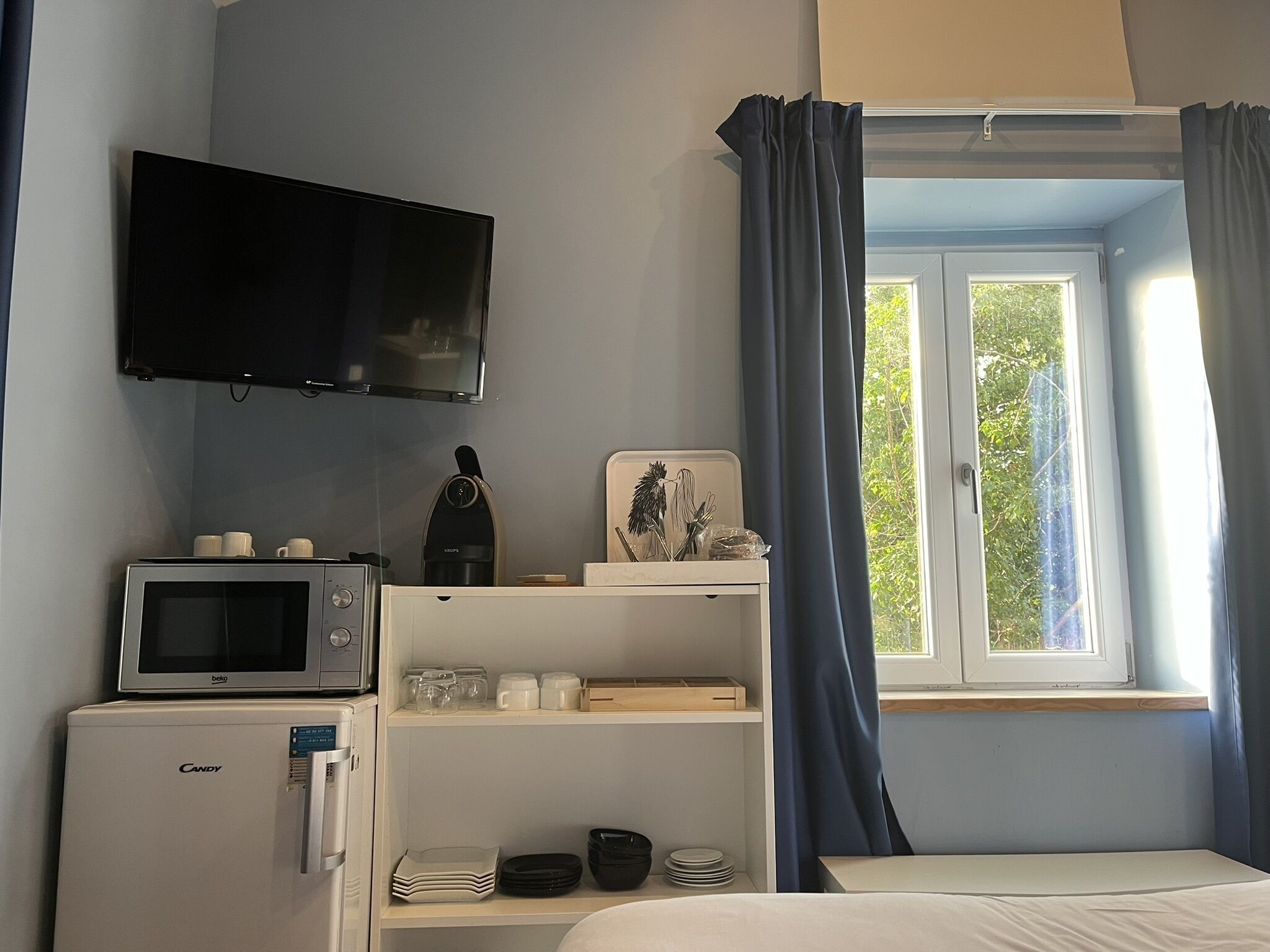 A hotel room with a bed, a television mounted on the wall, a microwave, a refrigerator, a window with curtains, and a shelf with various items.