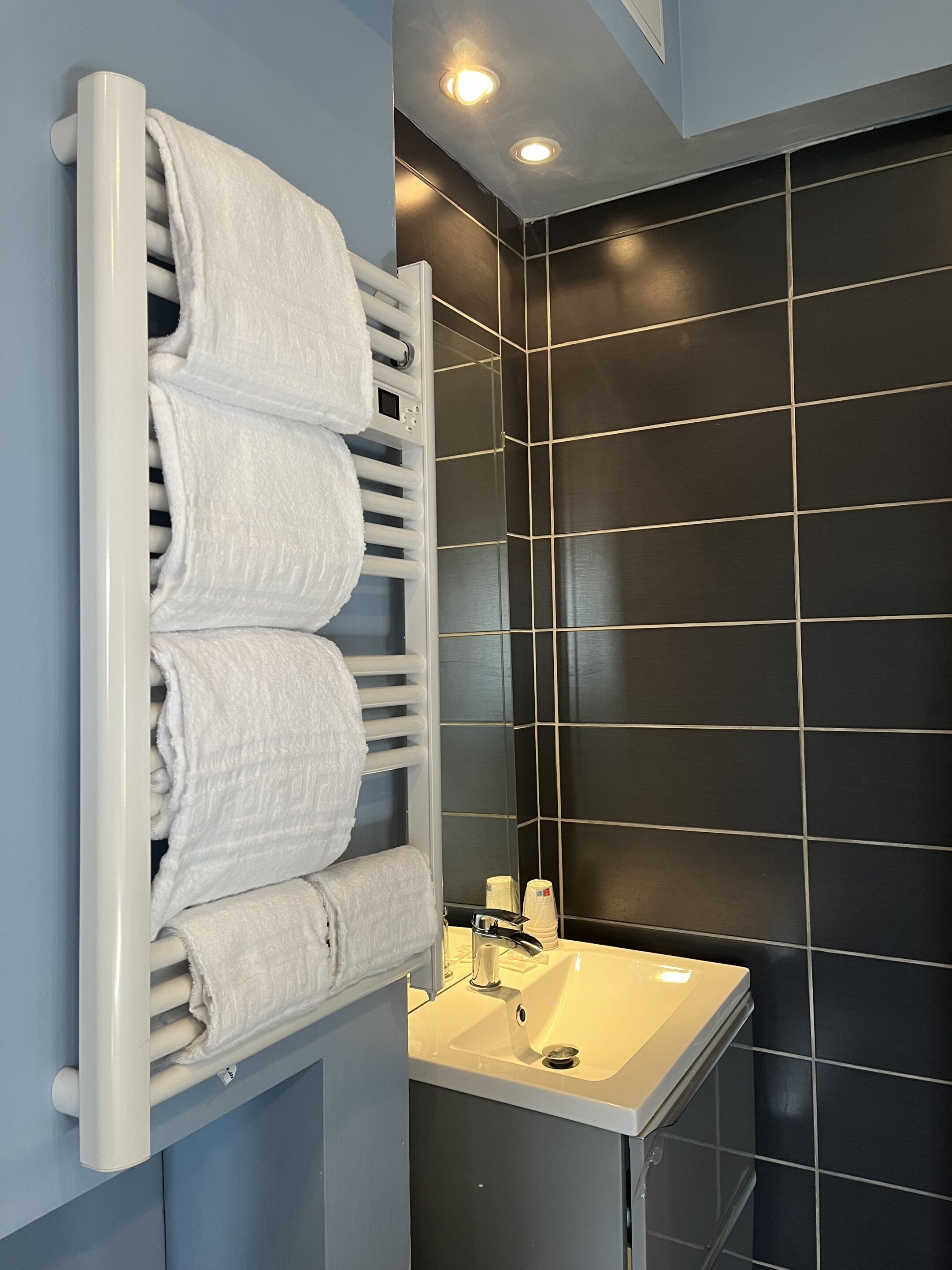 A bathroom with a white towel rack holding three towels, a white sink, and dark tiled walls.