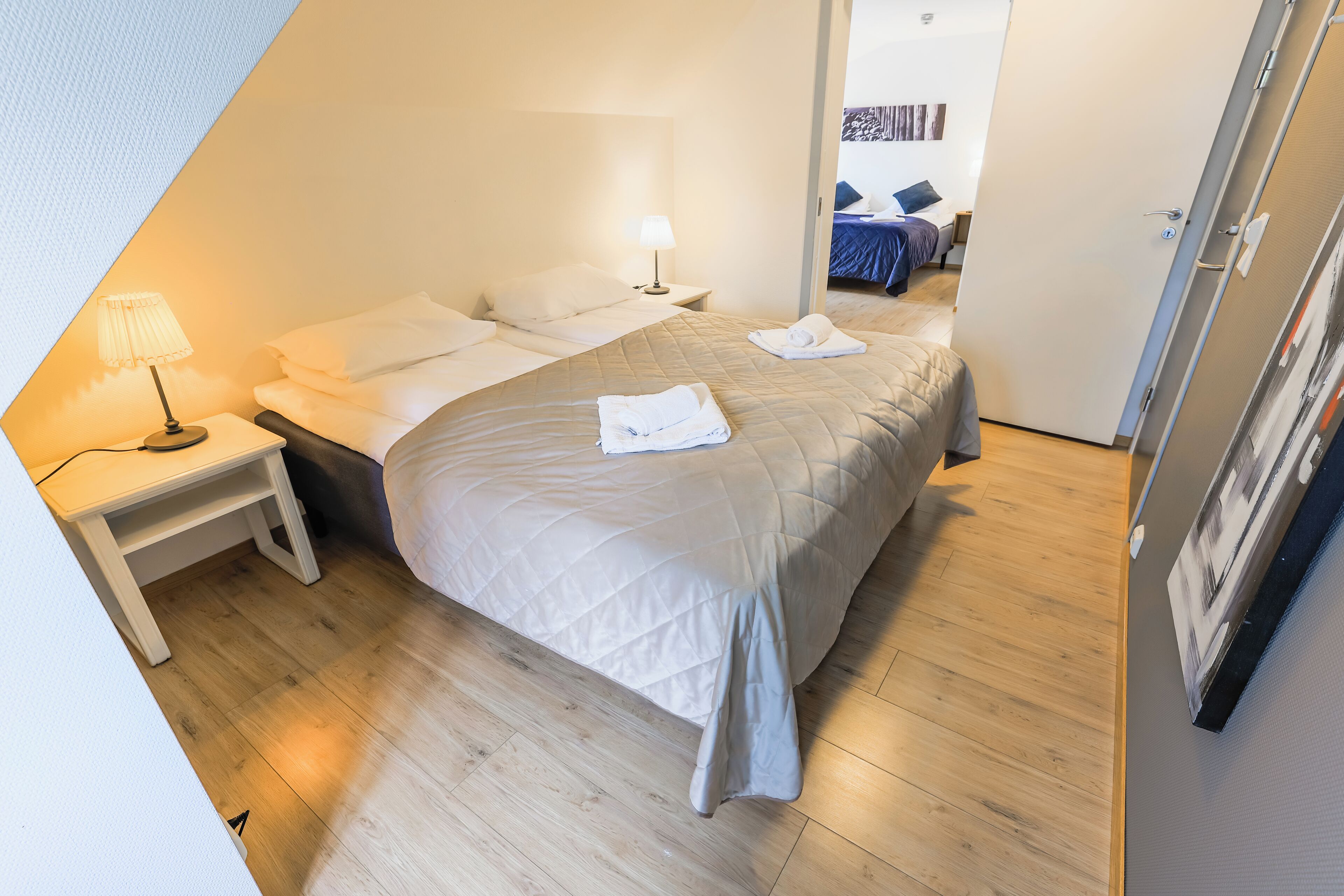 Een hotelkamer met een bed, nachtkastje, lamp en een deur naar een andere kamer.