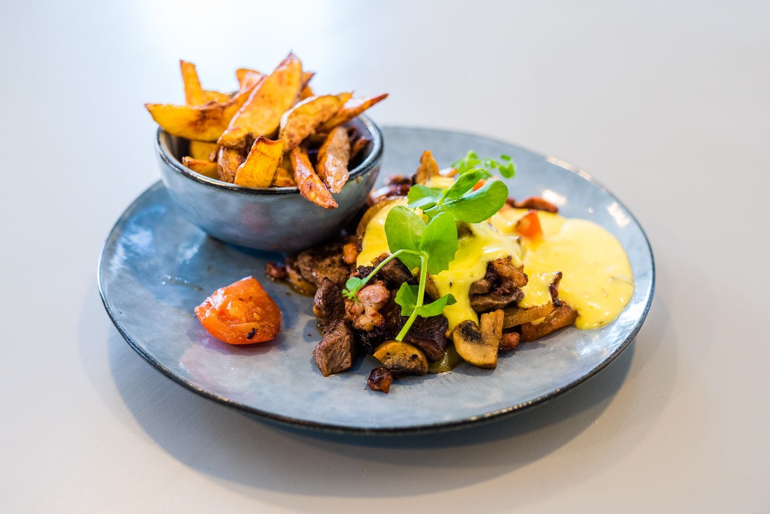 A plate with a steak topped with mushrooms and a creamy sauce, accompanied by a bowl of fries and a small tomato.