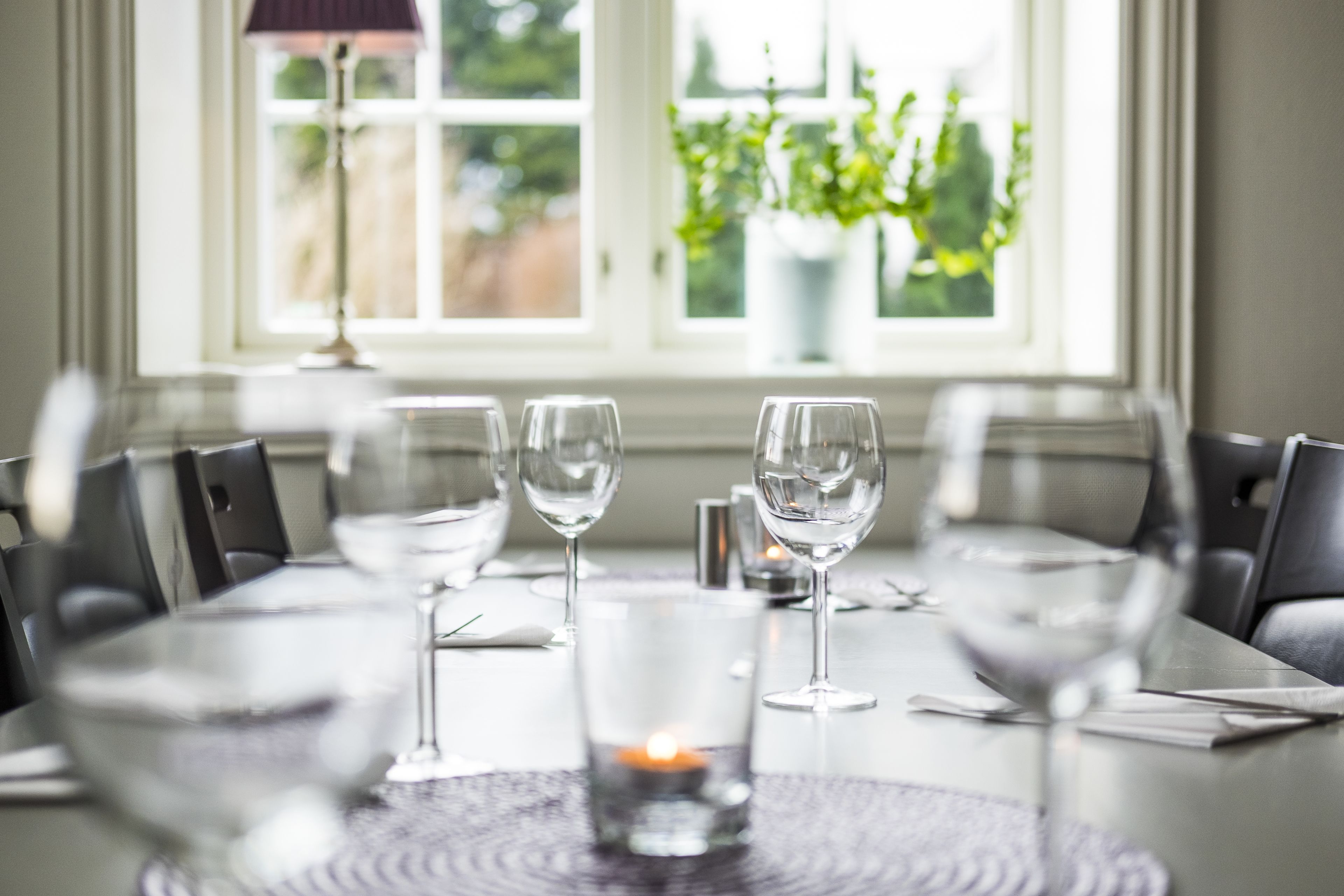 A dining table set for four with wine glasses and a candle, in a room with a view of greenery outside.