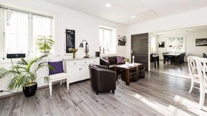 A spacious living area with a wooden floor, a white sofa, a dining table, and a coffee table. There are two potted plants and a chalkboard on the wall. - Verkshotellet (Strand)