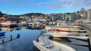 A marina with boats docked, a calm water surface, and a town with buildings in the background. - Verkshotellet (Strand)