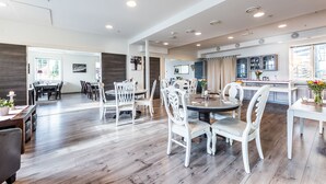 A spacious dining area with white upholstered chairs and a round table, a kitchen with modern appliances, and a large window providing natural light. - Verkshotellet (Strand)