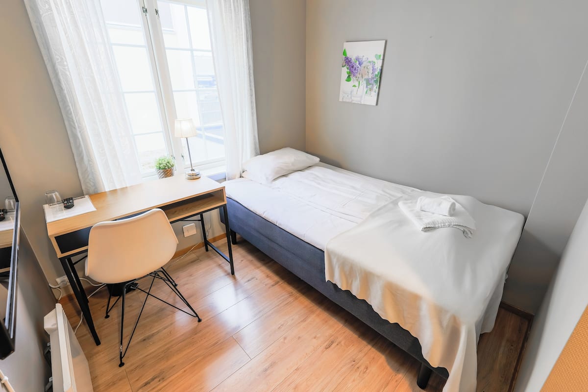 Ein kleines, ordentlich eingerichtetes Schlafzimmer mit einem Einzelbett, einem Schreibtisch, einem Stuhl und einem Fenster mit Vorhängen.