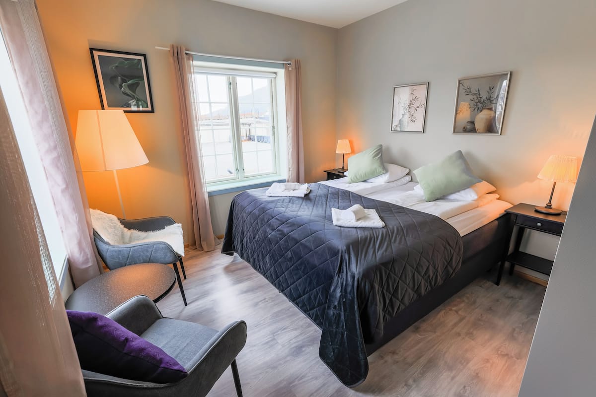 Ein Hotelzimmer mit Bett, zwei Nachttischen, einem Sessel, einem Fenster mit Vorhängen und Wandbildern.