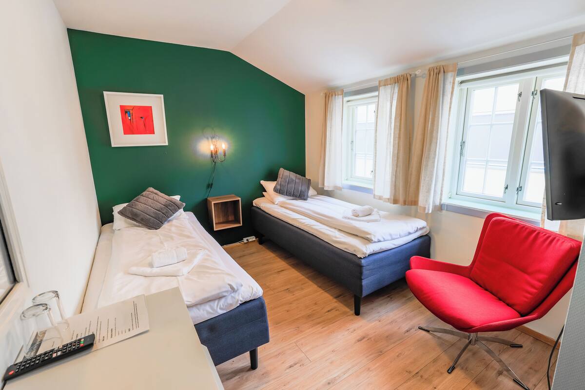 Ein Zimmer mit zwei Betten, einem roten Sessel, einem Fernseher und einem Fenster mit Vorhängen.
