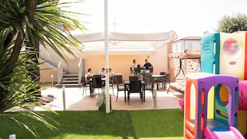 Uma área de pátio externa com uma mesa e cadeiras para refeições, um parquinho infantil e uma escadaria que leva a um andar superior.