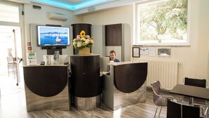 A reception area with a TV, a flower arrangement, and a window with a view of trees. - Hotel Roma (Massa)