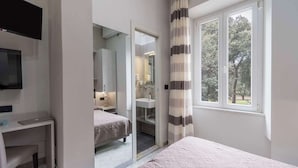 A bedroom with a bed, a desk, a TV, and a window with a view of trees. - Hotel Roma (Massa)