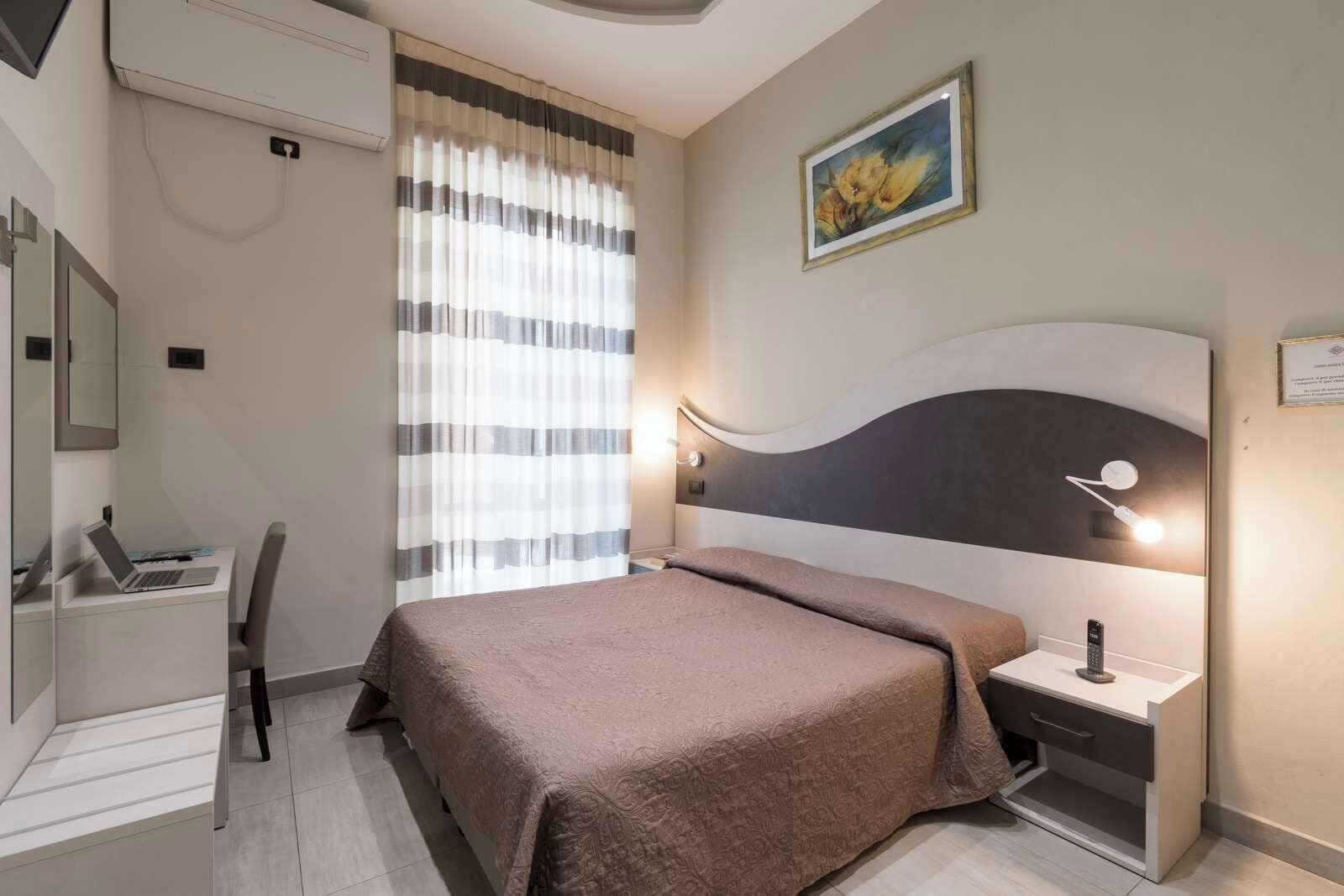 A hotel room with a bed, bedside table, lamp, and a desk with a laptop.