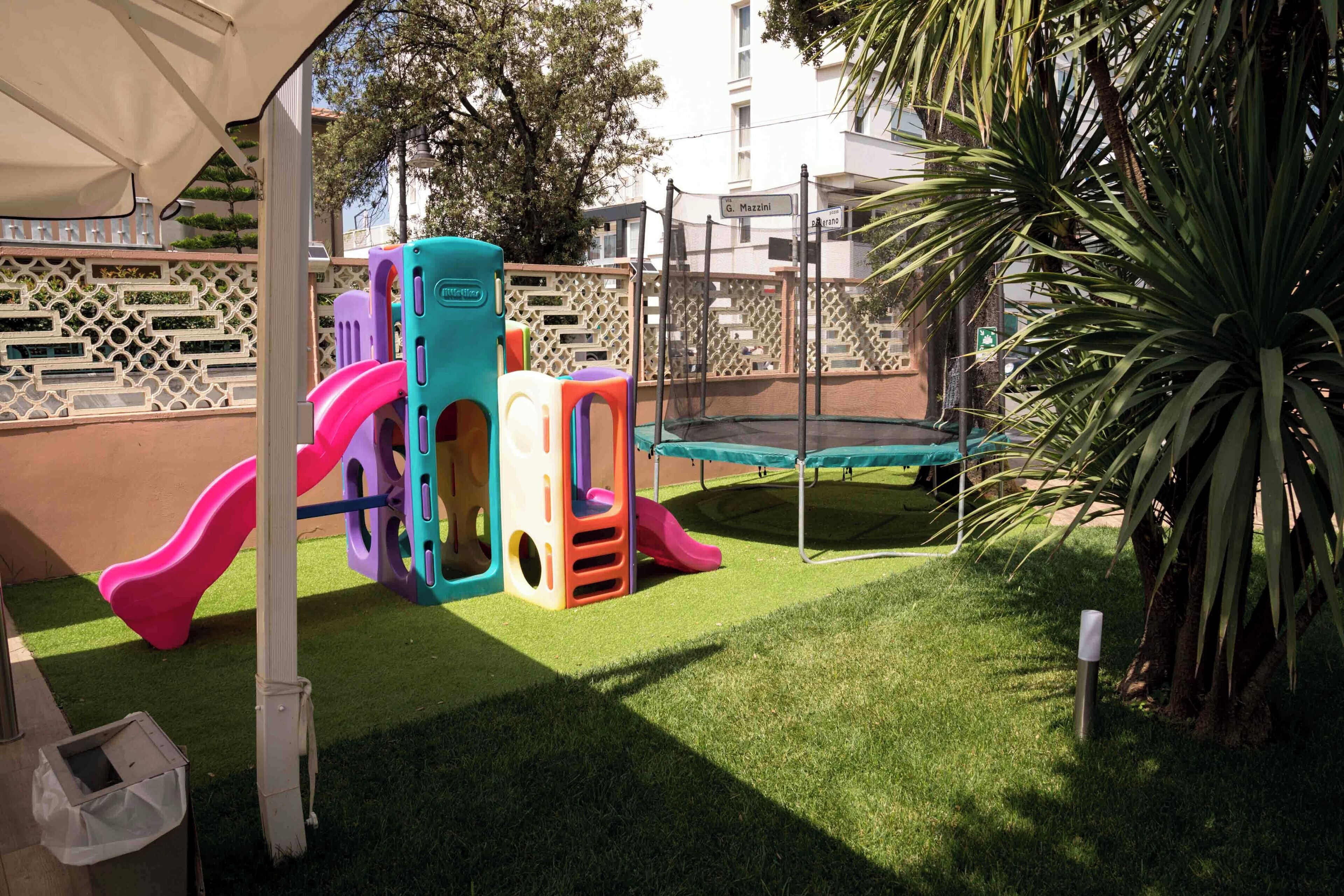 Un patio de juegos con toboganes y un trampolín, rodeado por una valla y una palmera.