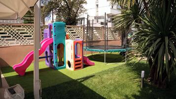 Un patio de juegos con toboganes y un trampolín, rodeado por una valla y una palmera.